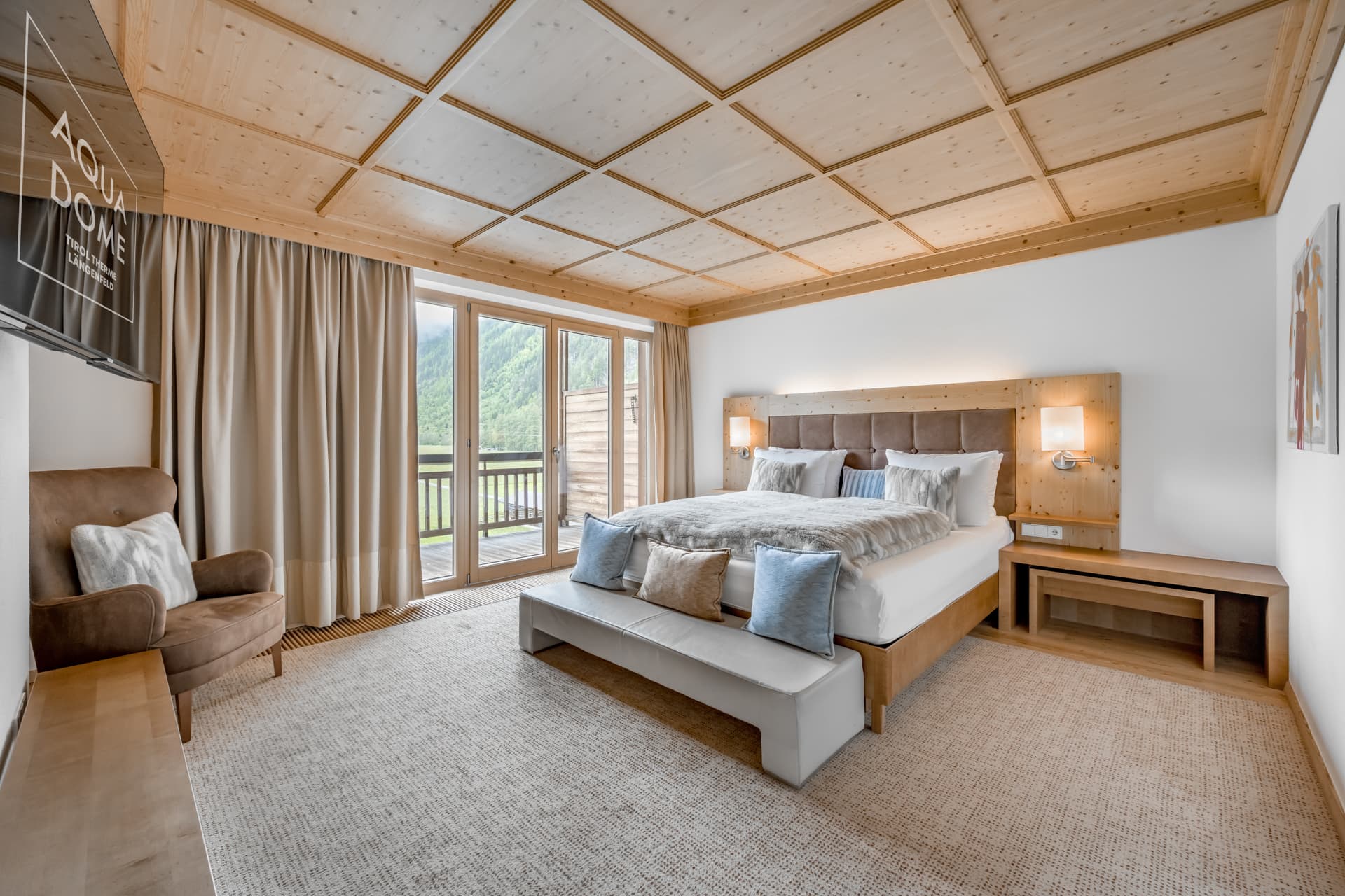 Современный гостиничный номер с большой кроватью, нейтральным декором, потолком из деревянных панелей, балконной дверью, креслом и видом на зеленые горы из окон.