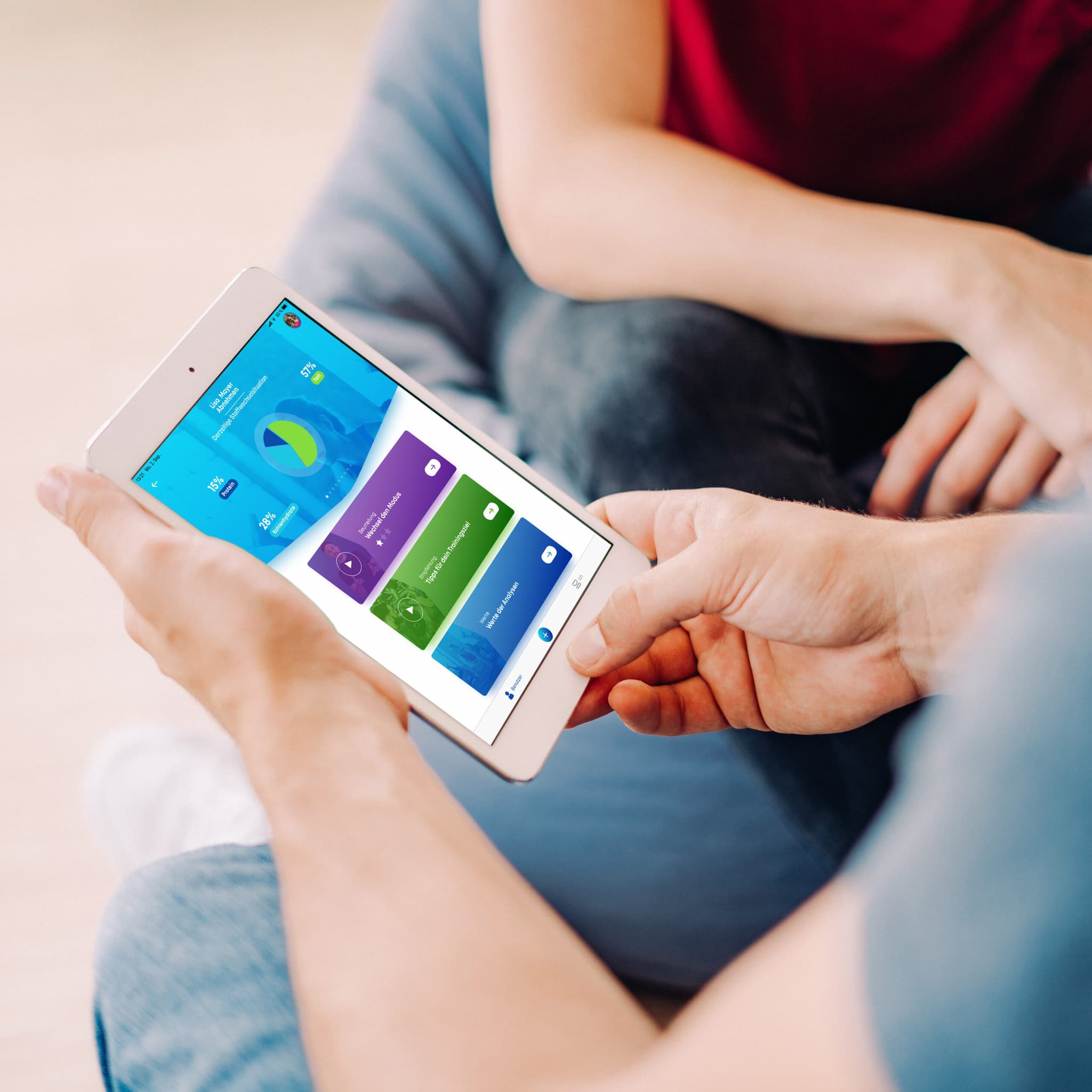 Eine Person hält ein Tablet in der Hand, auf dem eine bunte Finanz- oder Haushaltsplanungs-App angezeigt wird, während eine andere Person im Hintergrund sitzt.
