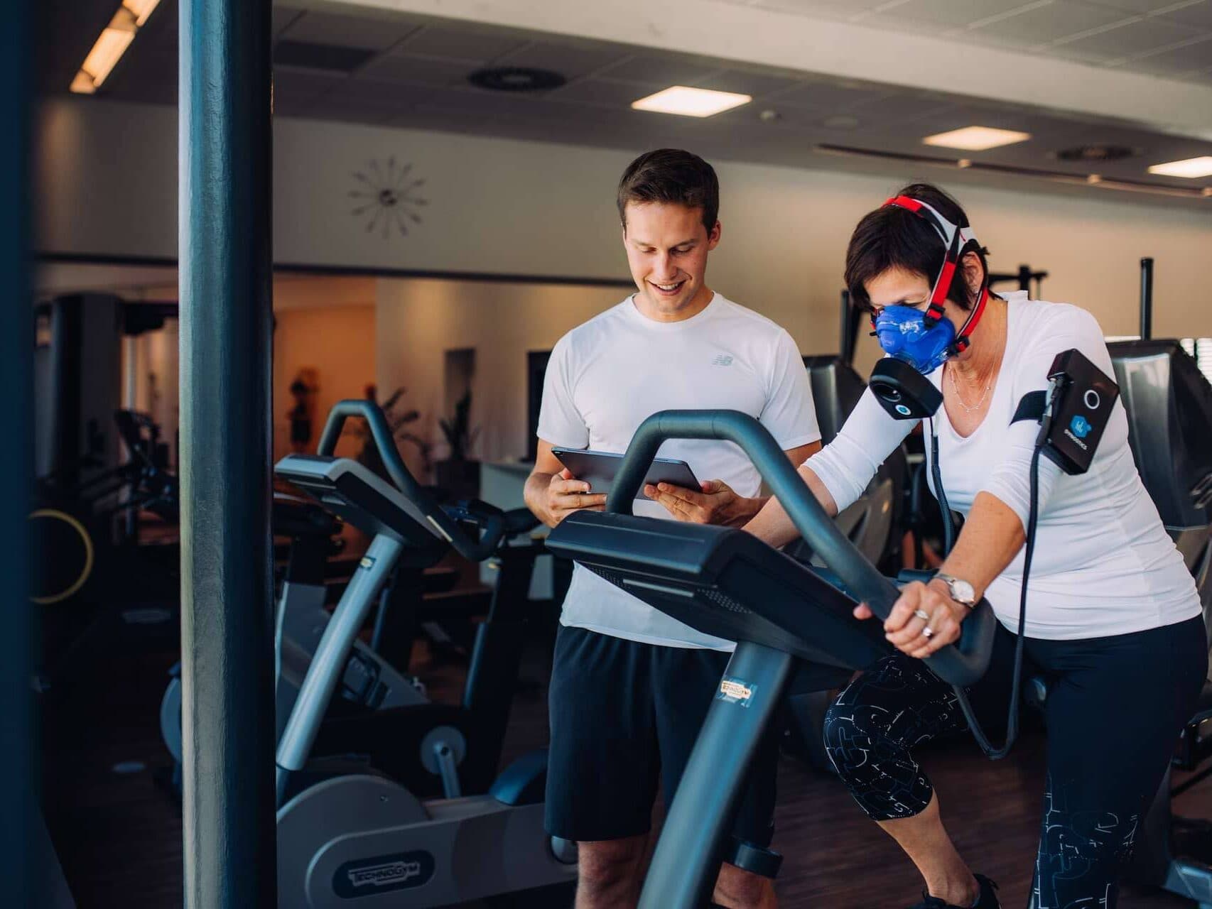 Eine Frau, die eine Fitnessmaske und Sensoren trägt, fährt auf einem stationären Fahrrad, während ein Trainer in einem Fitnessstudio die Daten auf einem Tablet überwacht.
