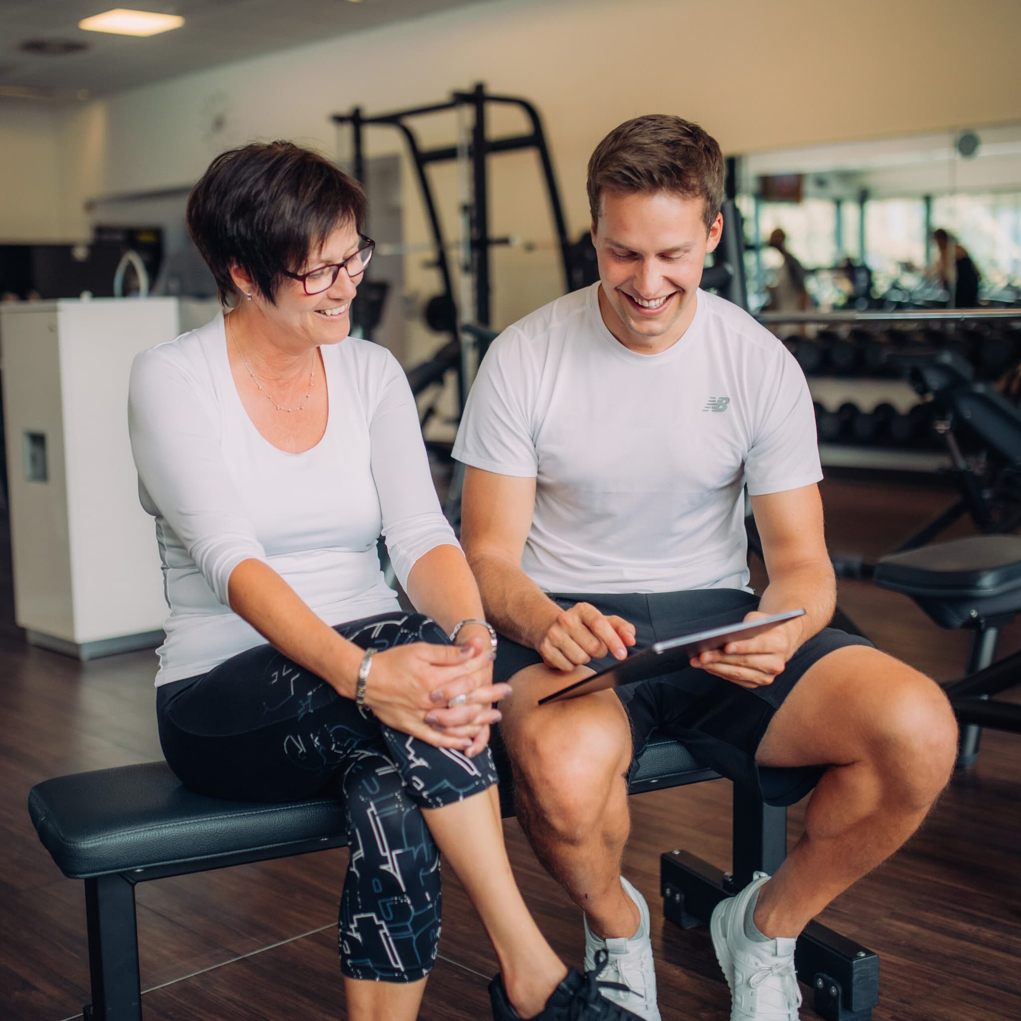 Eine Frau und ein Mann sitzen auf einer Bank in einem Fitnessstudio, schauen auf ein Tablet und lächeln. Im Hintergrund sind Trainingsgeräte zu sehen.