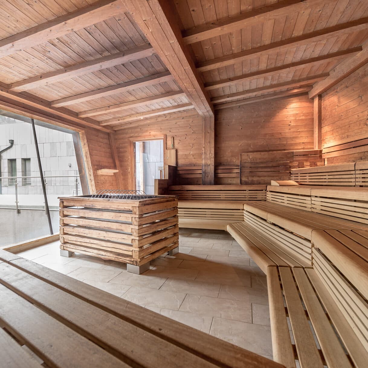 Geräumiger Saunaraum aus Holz mit Stufenbänken, großem Fenster und zentralem Saunaofen auf gefliestem Boden.