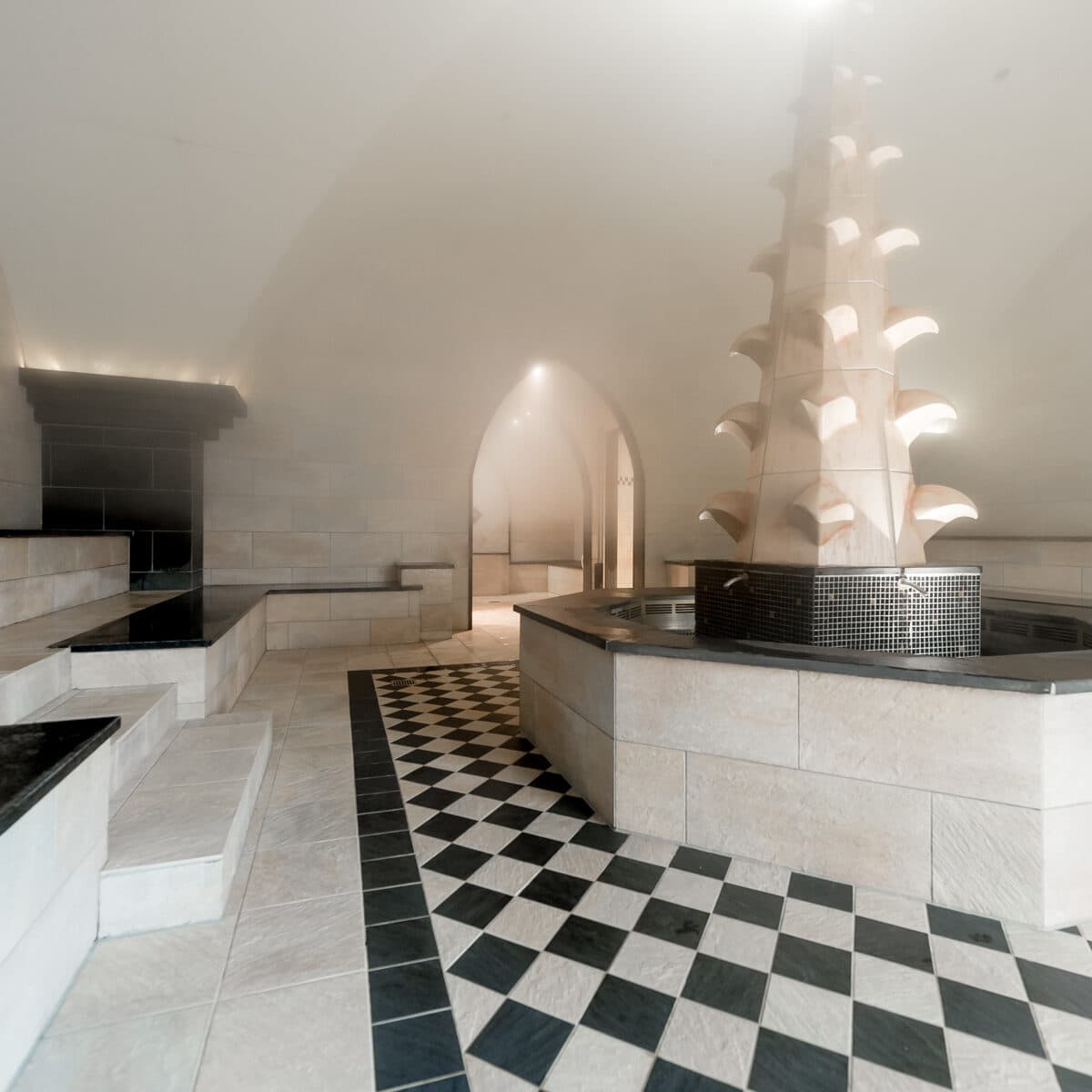 Mit Dampf gefüllter Spa-Raum mit gefliestem Boden, schwarz-weiß kariertem Weg, Steinbänken und einem zentralen dekorativen Steinbrunnen.