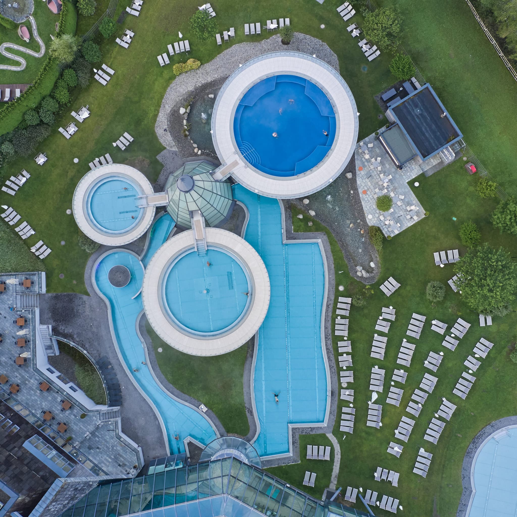 Luftaufnahme eines Freibadkomplexes mit kreisrunden Becken, Liegestühlen auf dem Rasen und umliegenden Landschaftsgärten.