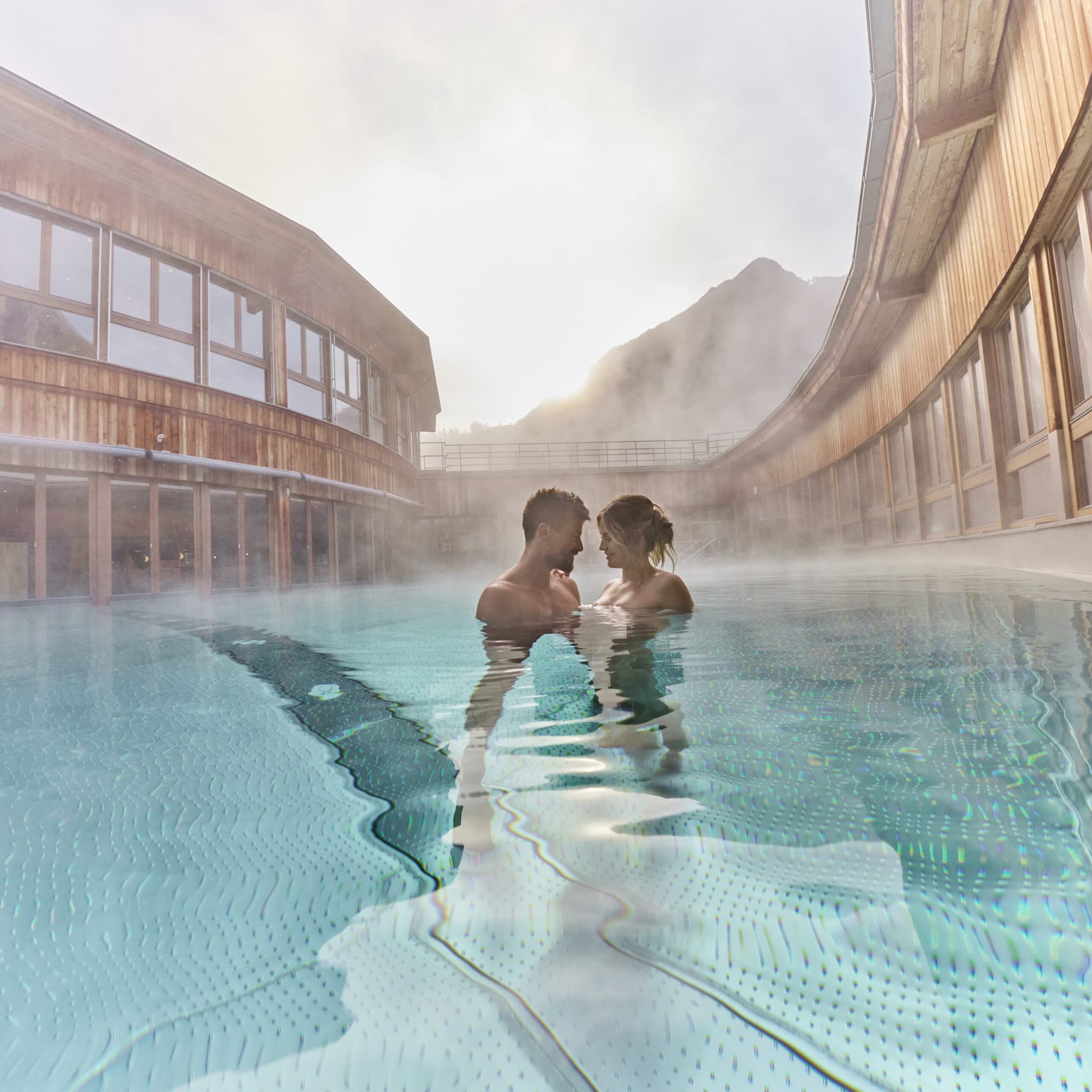 Ein Paar steht eng beieinander in einem dampfenden Freibad, umgeben von Holzhäusern und Bergen im Hintergrund.