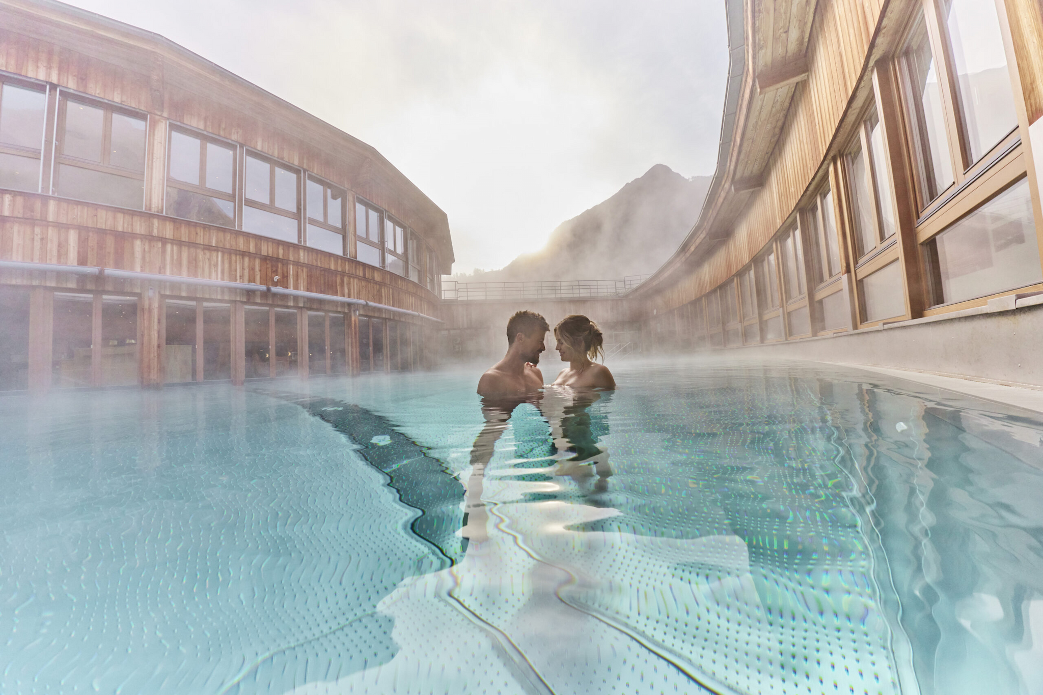 Una coppia è vicina in una piscina riscaldata all'aperto circondata da edifici in legno, con la nebbia che sale dall'acqua e le montagne visibili sullo sfondo.