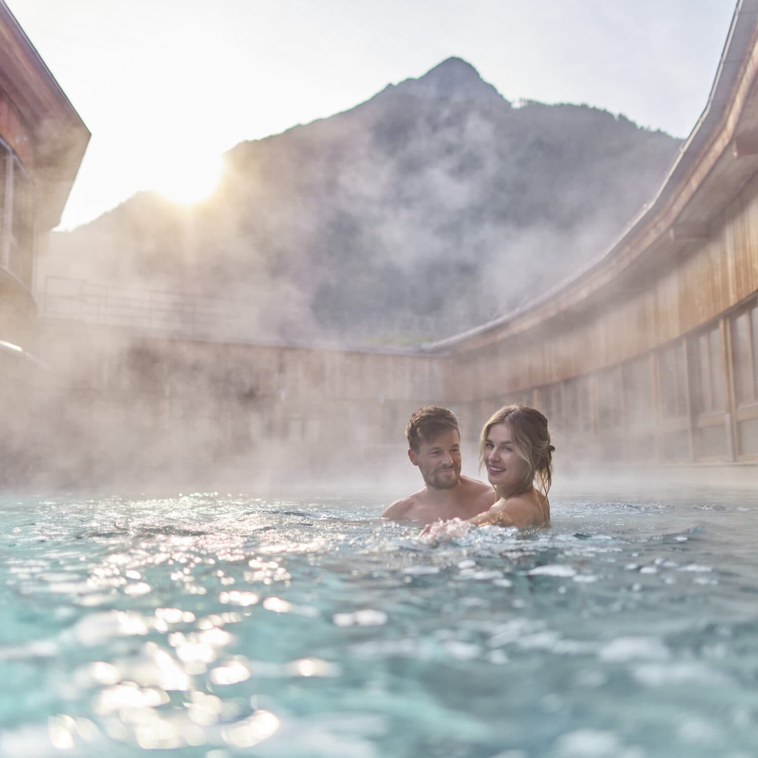 A couple enjoys an outdoor hot spring pool surrounded by steam, with wooden buildings and a mountain visible in the background.