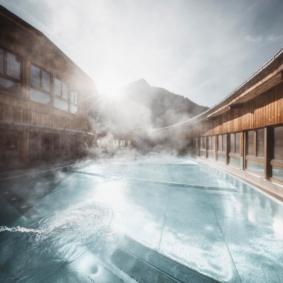 Outdoor swimming pool with steam rising, surrounded by wooden buildings, with sunlight streaming over a mountain in the background.