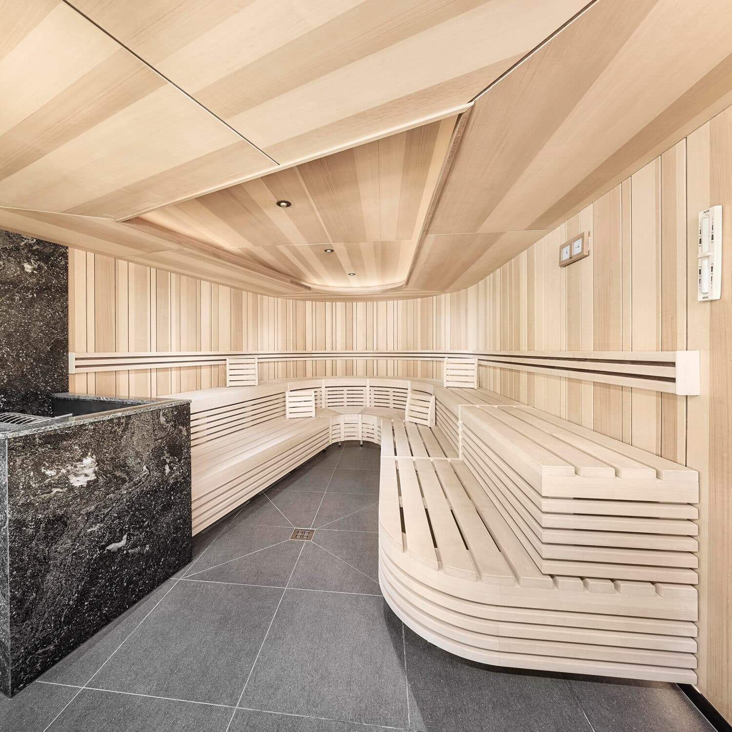Modern sauna with light wood benches, wood-paneled walls and ceiling, gray tile flooring, and a black stone heater in the corner.