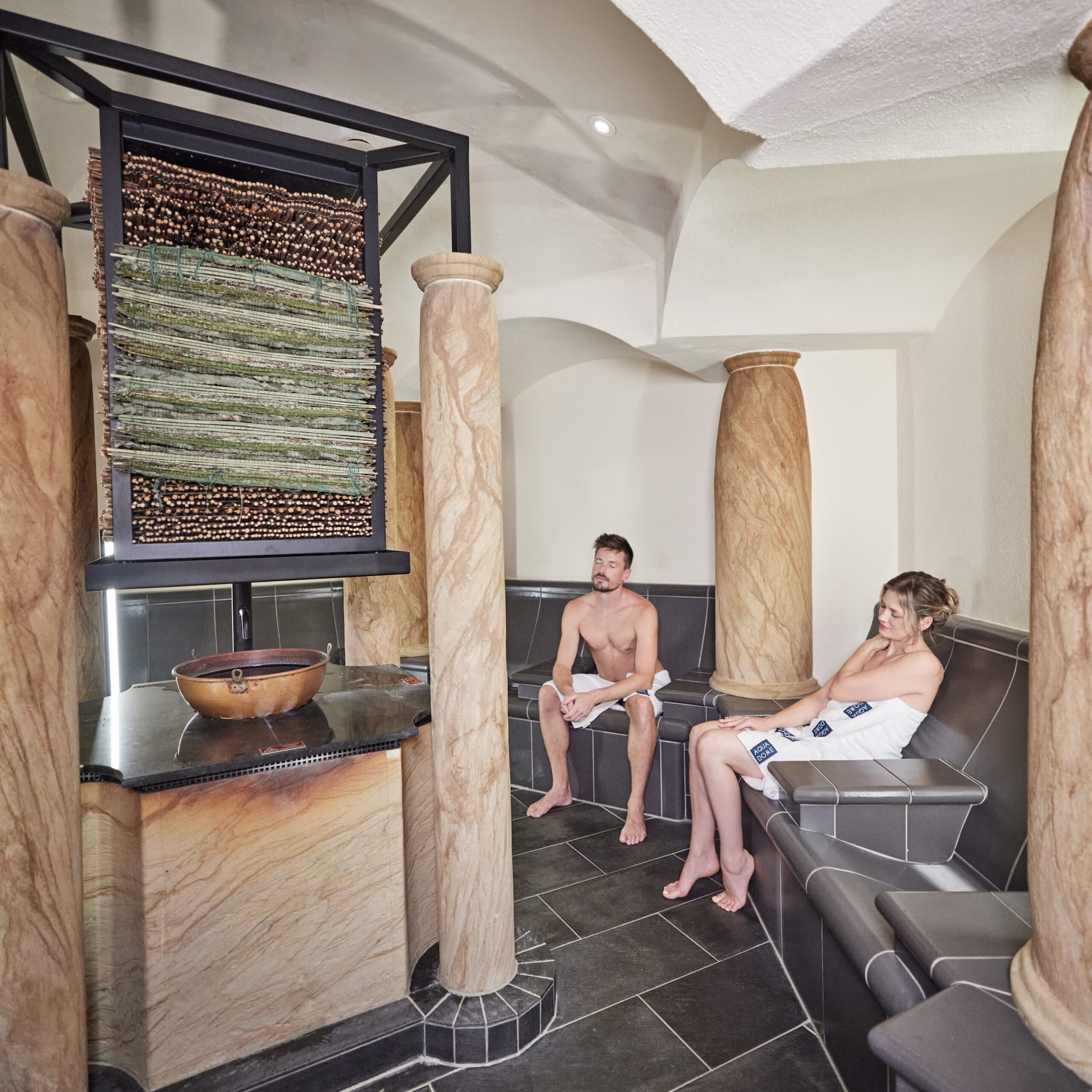 Due persone in asciugamano siedono su panche in una sala termale piastrellata con grandi colonne di pietra e un elemento decorativo centrale.