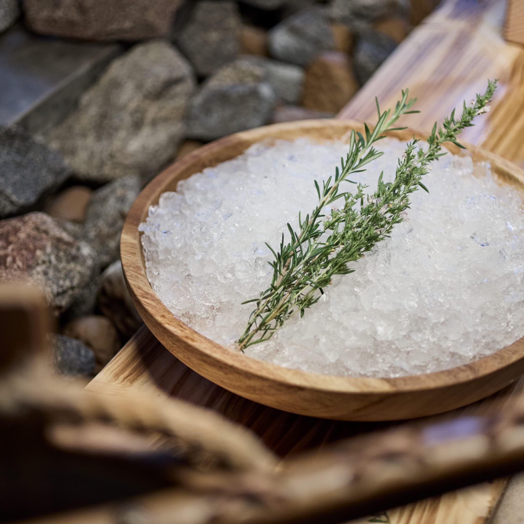 Una ciotola di legno riempita di ghiaccio tritato e guarnita con due rametti di erbe fresche, posta su un piano di legno con pietre sullo sfondo.