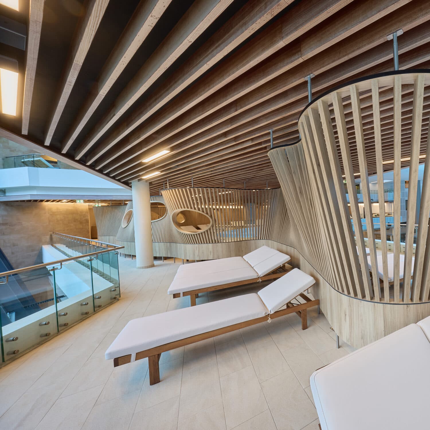 Modern indoor lounge area with wooden slatted walls and ceiling, featuring white cushioned lounge chairs and glass railing overlooking another level.
