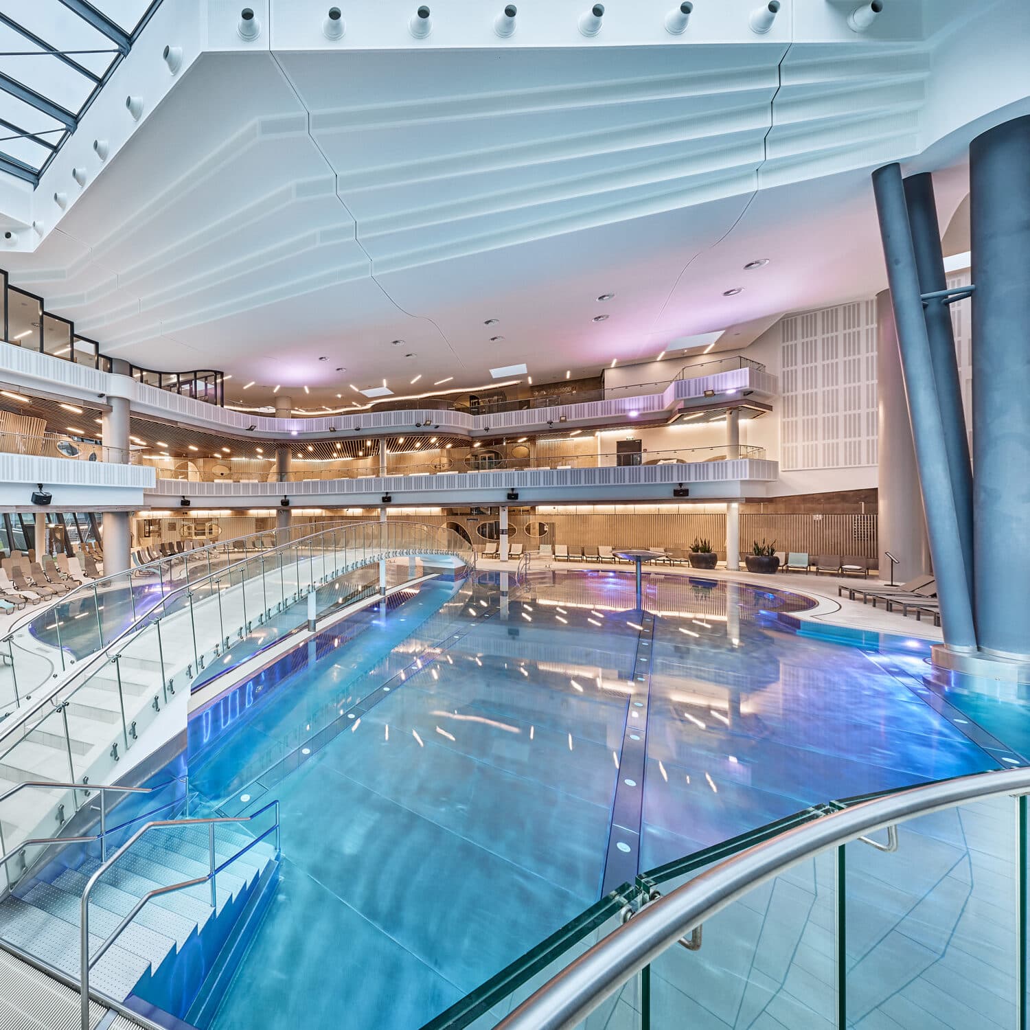 Indoor swimming pool in a modern, multi-level facility with glass railings, overhead lighting, and several balconies overlooking the water.