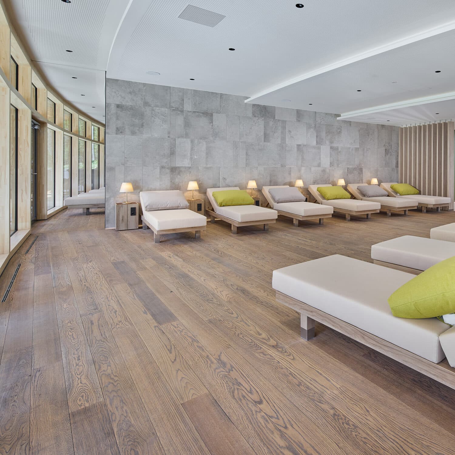 Spacious relaxation room with wooden floors, neutral lounge chairs, green pillows, and small lamps, set against a stone accent wall and large windows on one side.