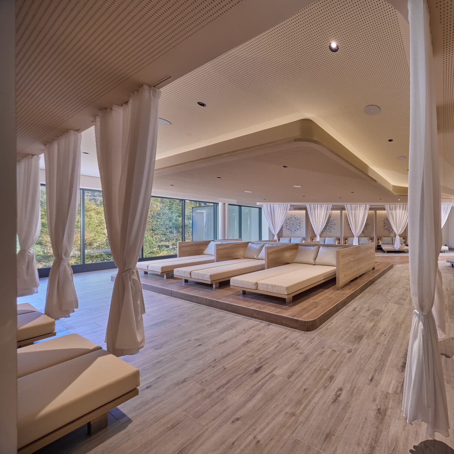 A spacious, modern relaxation lounge with large cushioned beds, white curtains, wood flooring, and floor-to-ceiling windows overlooking greenery.