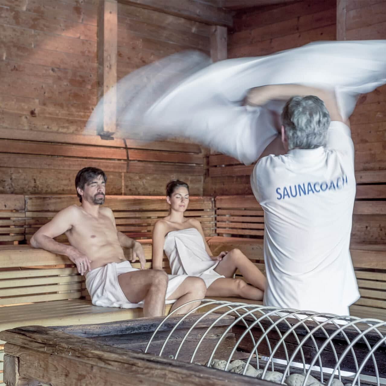 Ein Mann und eine Frau sitzen in Handtücher gehüllt in einer Holzsauna, während eine Person, die ein SAUNACOACH-Shirt trägt, mit einem Handtuch wedelt, um die Luft zu verteilen.