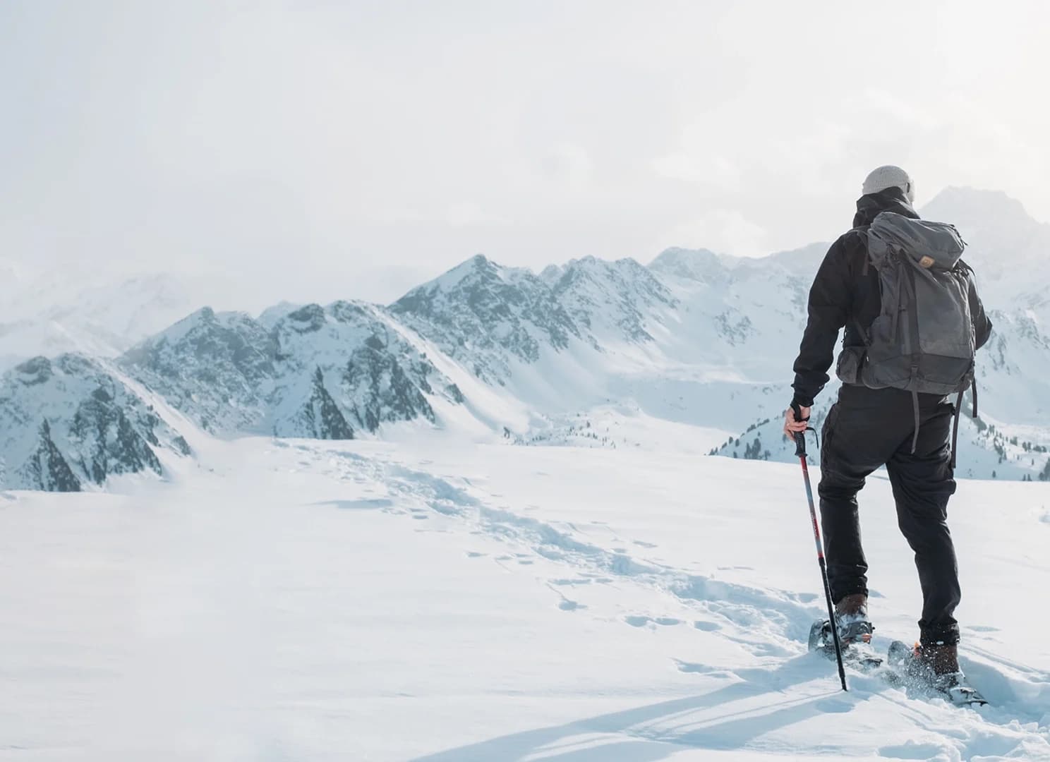 Eine Person in Winterkleidung mit Rucksack und Trekkingstock geht durch den Schnee auf ferne Berge unter einem bewölkten Himmel zu.