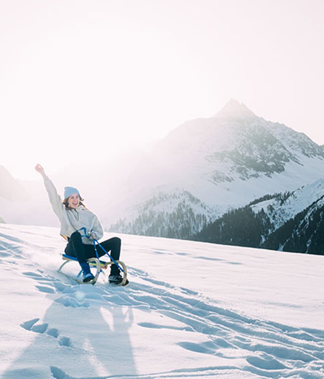 Eine Person, die auf einem Schlitten einen verschneiten Hügel hinunterfährt, mit Bergen im Hintergrund, und dabei einen Arm in die Luft hebt, im hellen Sonnenlicht.