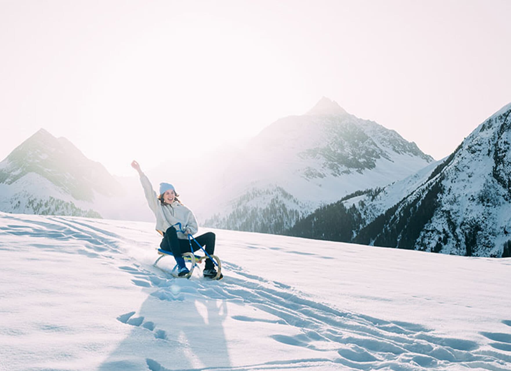 Eine Person fährt mit erhobenem Arm auf einem Schlitten einen verschneiten Hang hinunter, umgeben von schneebedeckten Bergen unter einem klaren Himmel.