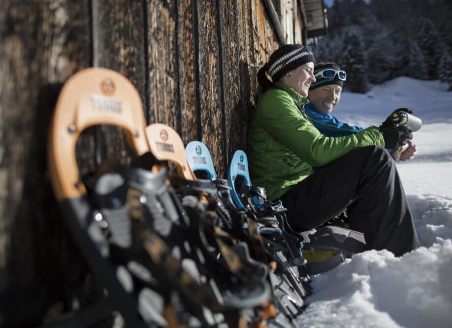Zwei Personen in Winterkleidung sitzen an einer Holzwand im Schnee, vor ihnen sind mehrere Paar Schneeschuhe aufgereiht.