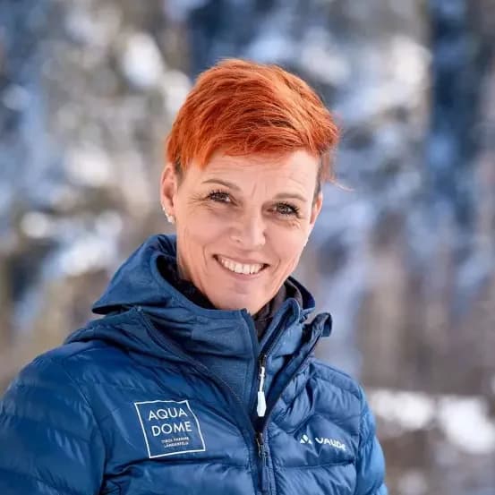 Eine Person mit kurzen roten Haaren und einer blauen Winterjacke steht im Freien vor einem verschneiten, unscharfen Hintergrund.