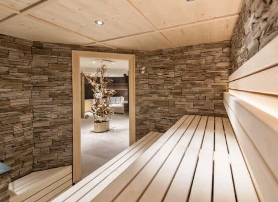 Ein moderner Saunaraum mit Holzbänken und Steinwänden, der durch eine offene Tür zu einem Lounge-Bereich mit einer dekorativen Holzskulptur führt.