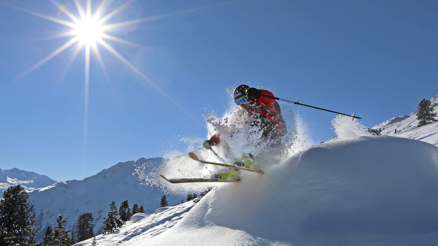Ein Skifahrer in roter Ausrüstung springt über einen schneebedeckten Abhang, wobei er eine Gischt aus Pulverschnee erzeugt, mit Bergen und einer hellen Sonne im Hintergrund.