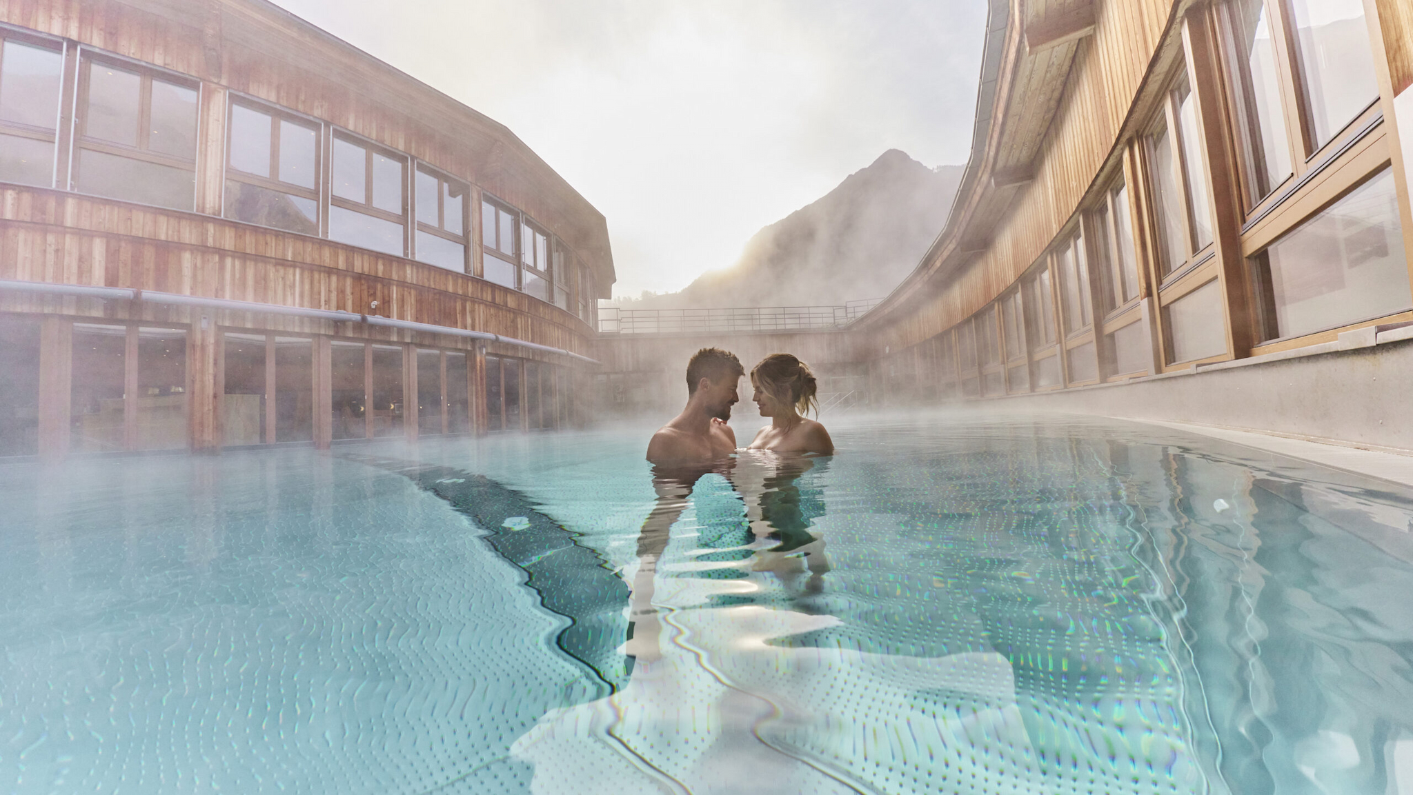 Deux personnes se tiennent l'une à côté de l'autre dans une piscine d'eau chaude en plein air, entourée de bâtiments en bois, avec de la brume qui s'élève et une montagne visible en arrière-plan.