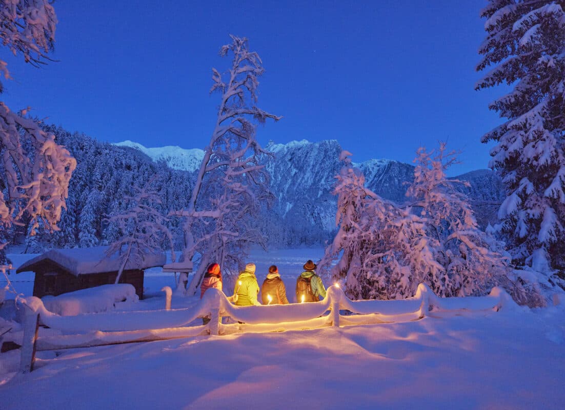 Drei Personen in Winterkleidung stehen in der Abenddämmerung an einem verschneiten Zaun und halten Laternen, umgeben von schneebedeckten Bäumen und Bergen im Hintergrund.