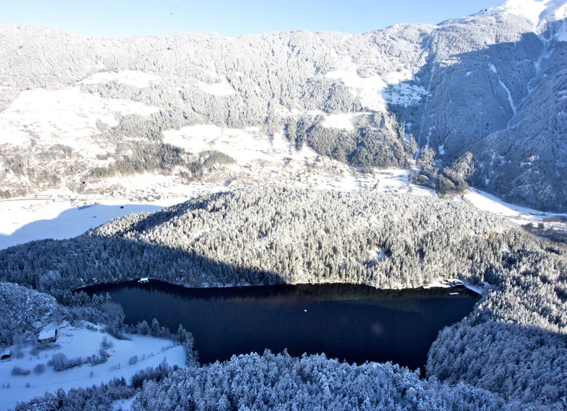 Eine schneebedeckte Landschaft mit einem dunklen, von Wäldern umgebenen See am Fuße der sonnenbeschienenen Berge.