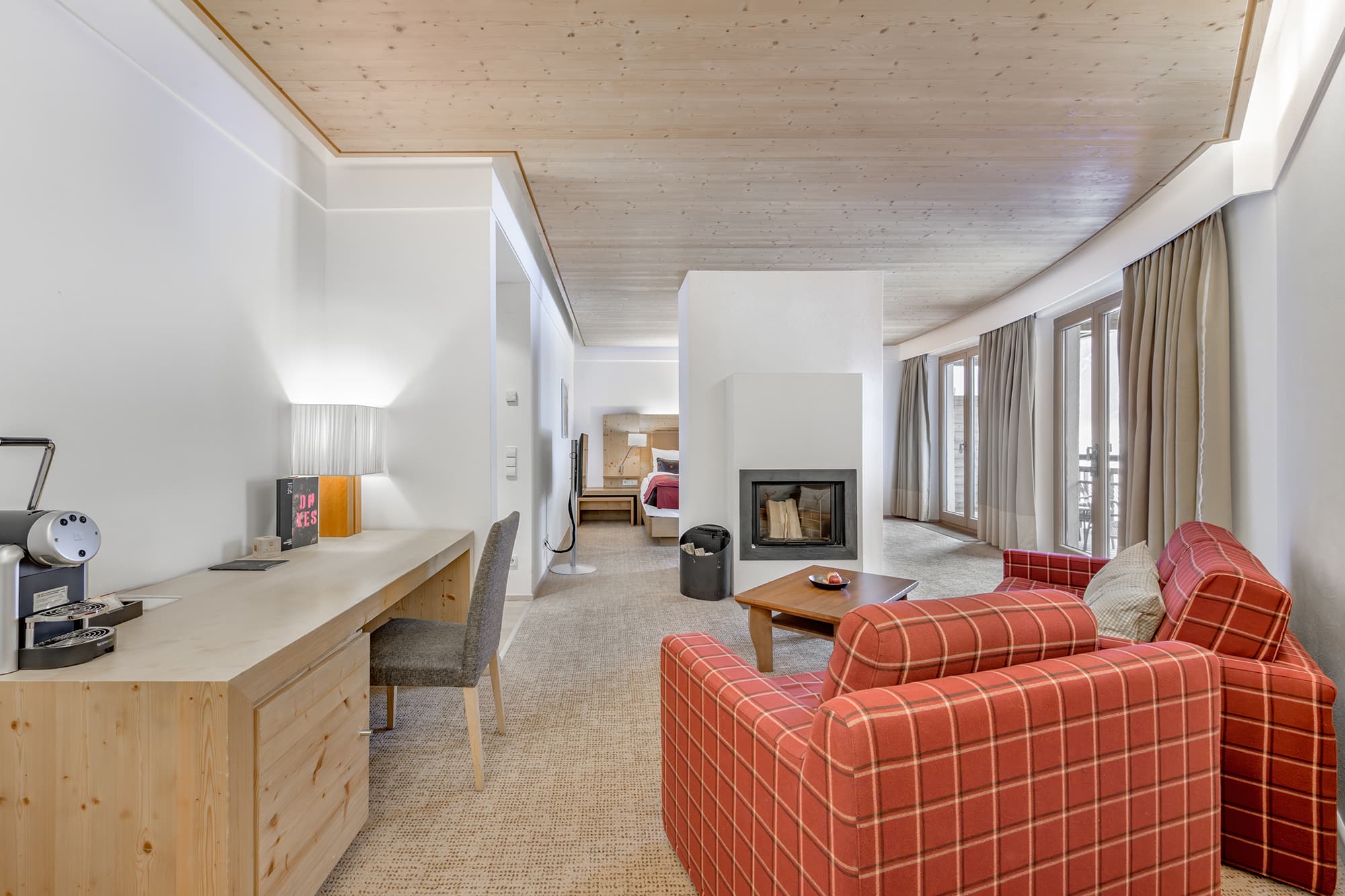 Spacious hotel suite with a work desk, red plaid armchairs, a fireplace, a bed in the background, and large windows with beige curtains.