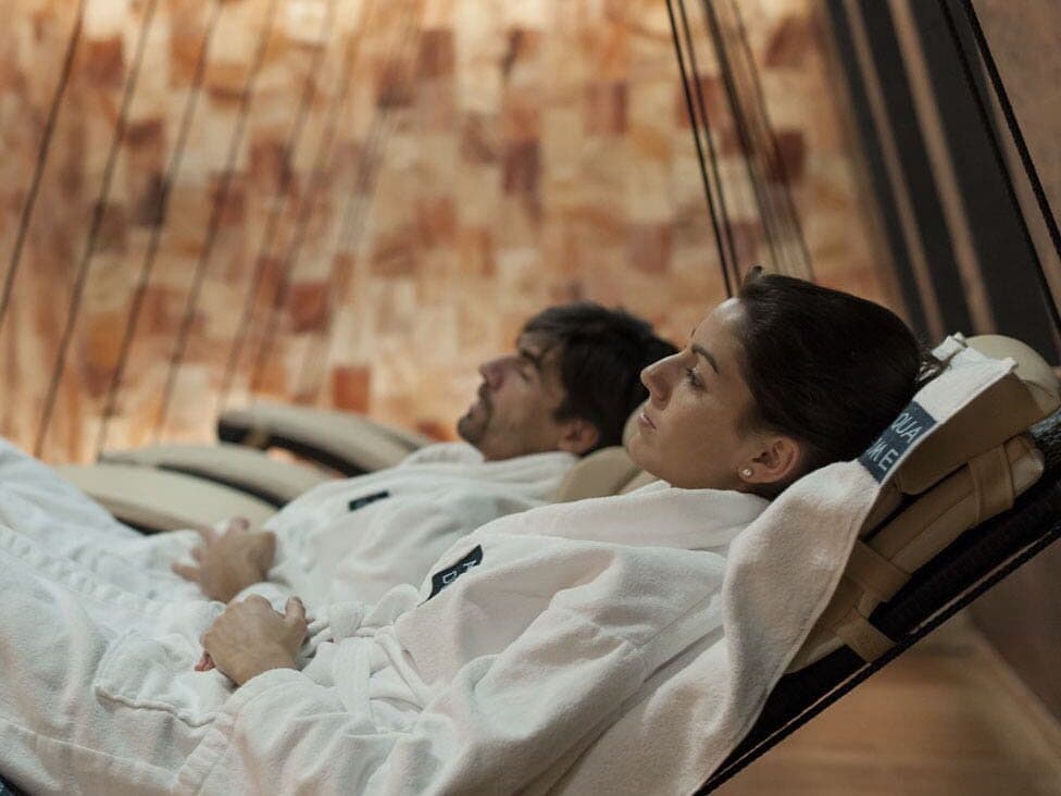 Zwei Personen in weißen Bademänteln entspannen sich auf Liegestühlen in einer spaähnlichen Umgebung mit einer warmtonigen, strukturierten Wand im Hintergrund.