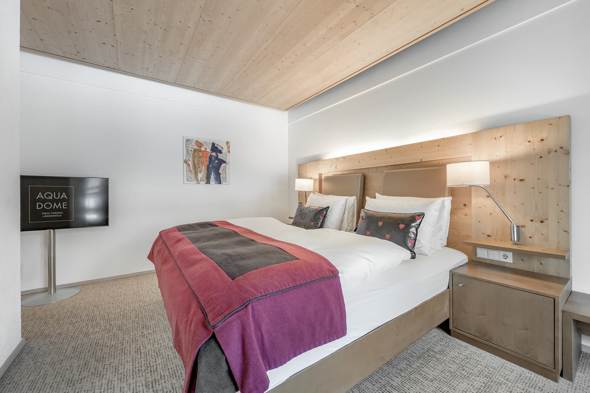 Modern hotel room with a double bed, wooden headboard, wall-mounted lamps, TV on a stand, and a painting on a white wall. The bed is covered with a red and gray blanket.