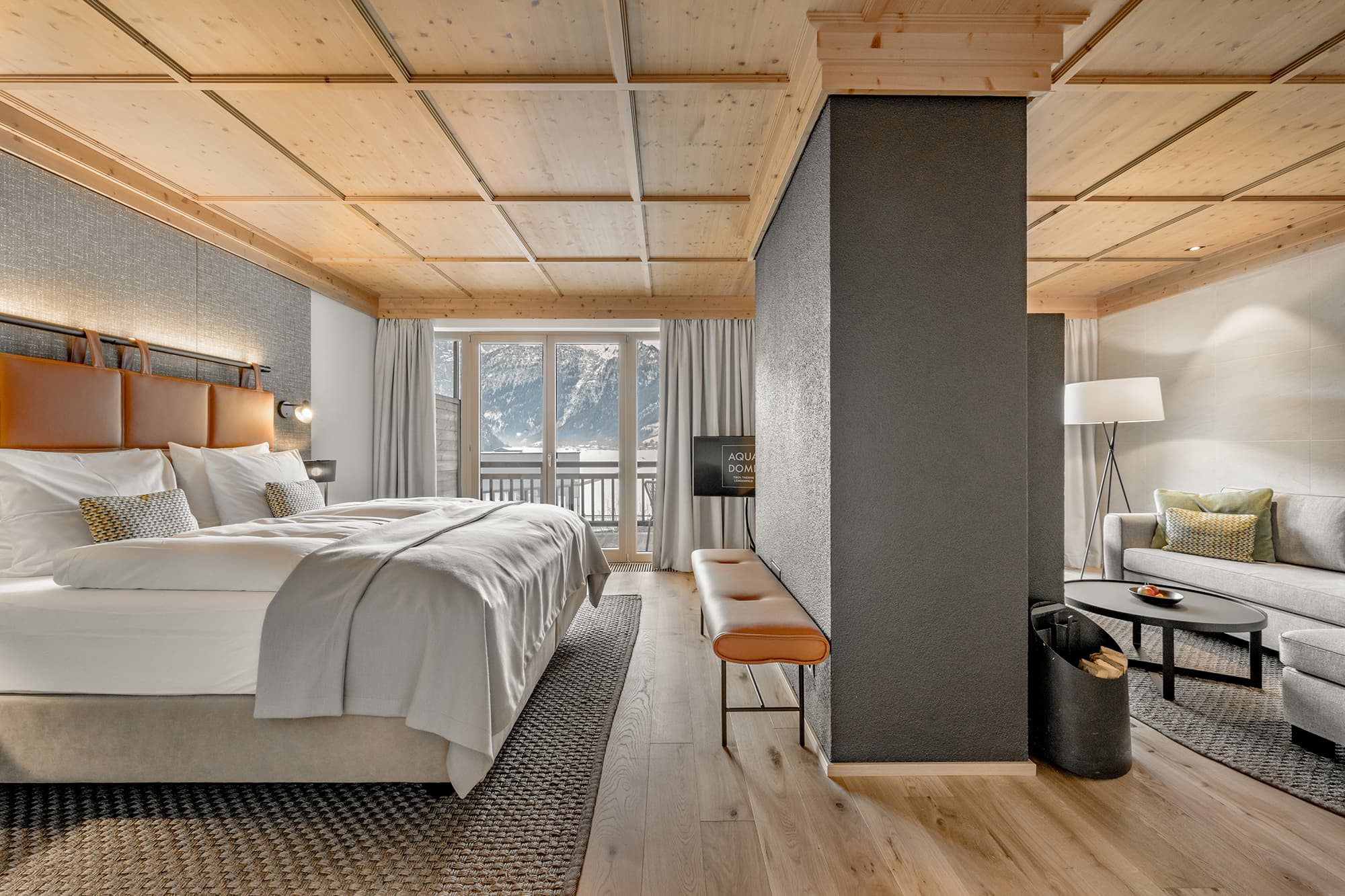 Modern hotel room with a large bed, wooden ceiling, gray accent wall, sitting area with a sofa, and a window overlooking mountains.