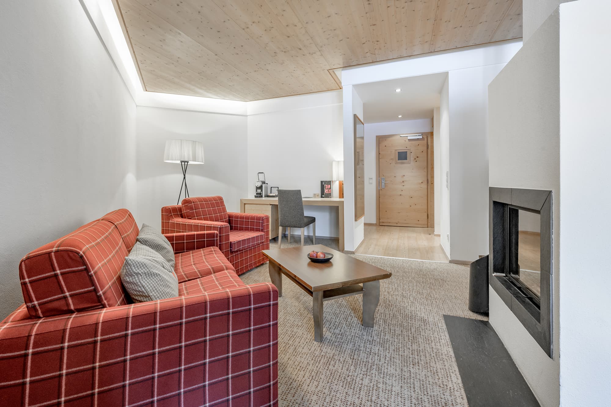 Modern hotel living area with two red plaid armchairs, a wooden coffee table, desk, floor lamp, fireplace, and a wooden entry door in the background.