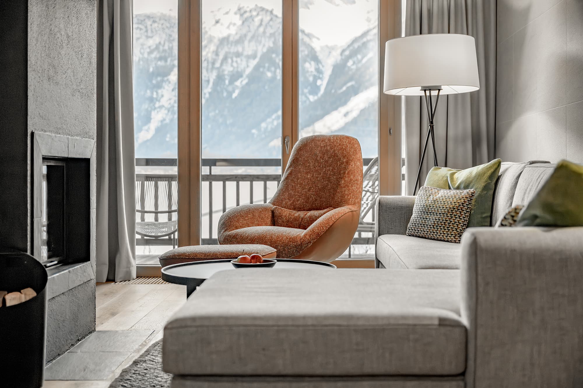A modern living room with a gray sectional sofa, an orange lounge chair, a floor lamp, and large windows overlooking snowy mountains.