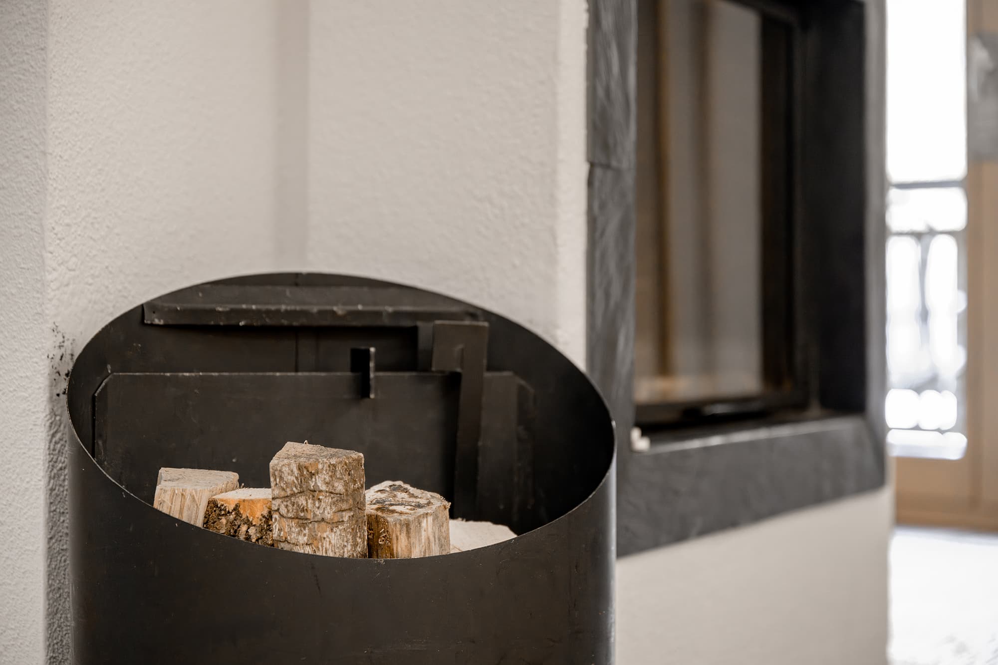 A black metal container filled with firewood sits against a white wall near a window.