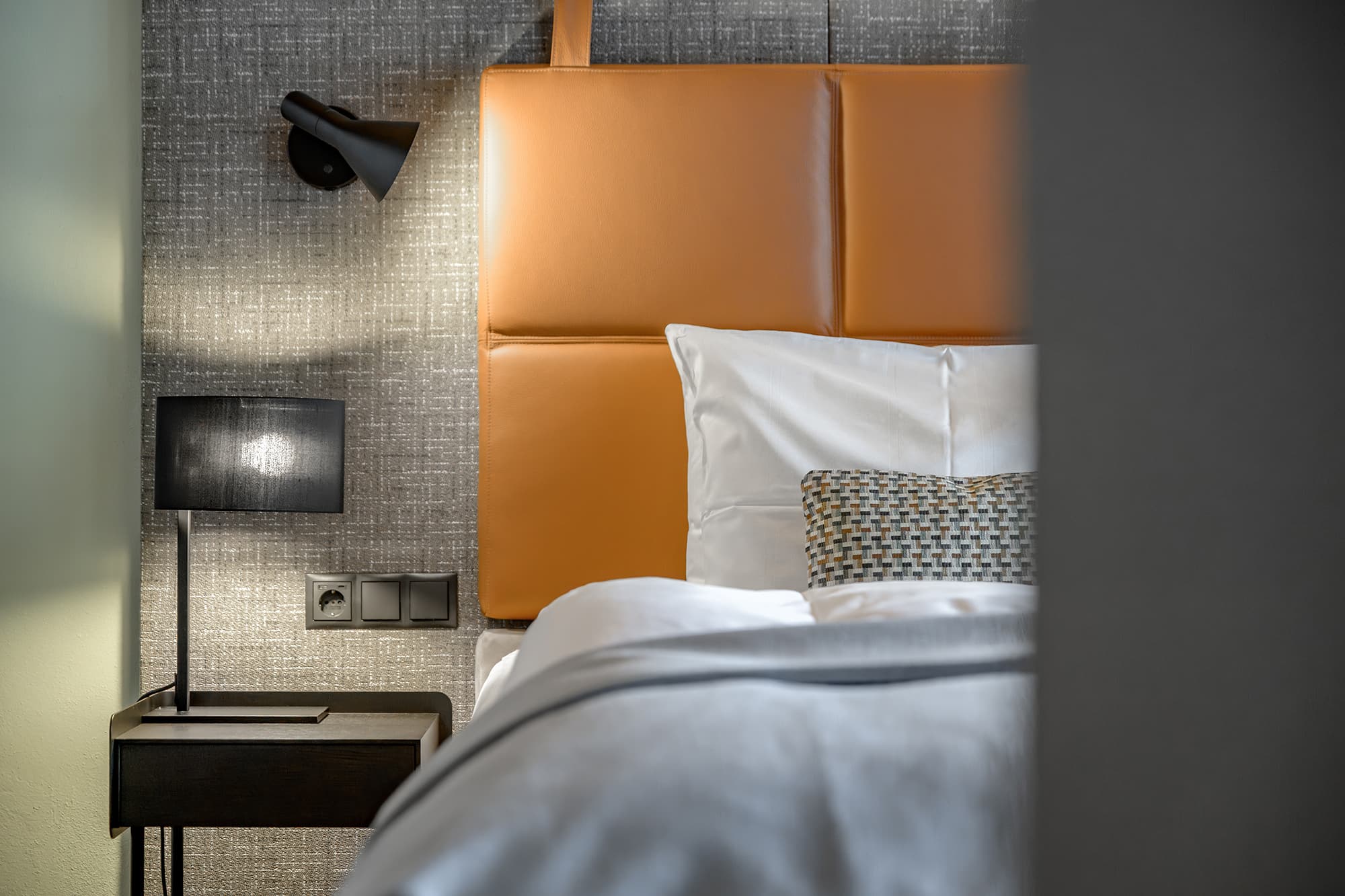 A modern hotel room features a bed with an orange cushioned headboard, white bedding, a patterned pillow, a small bedside table, and wall-mounted and table lamps.
