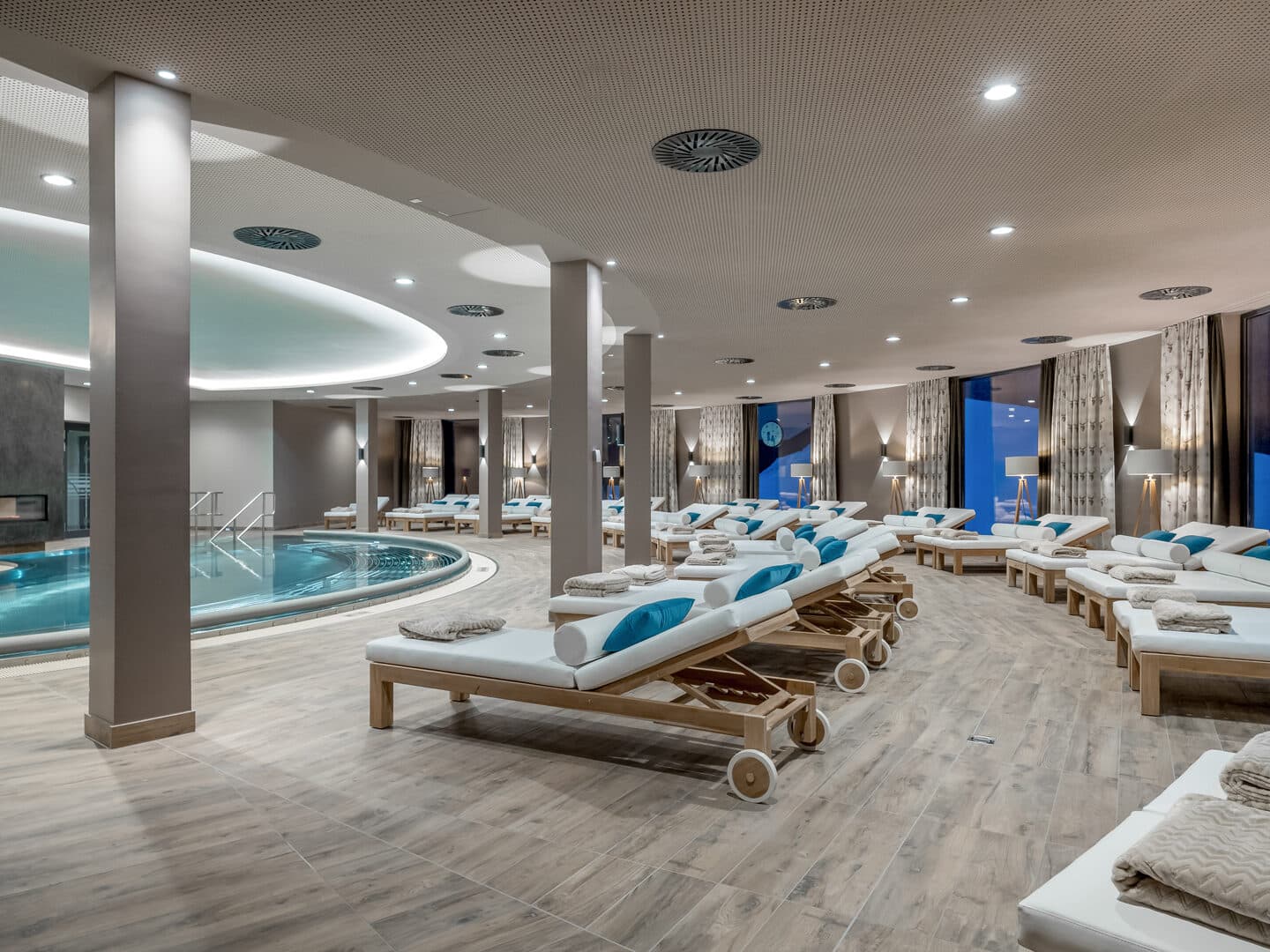 Modernes Indoor-Spa mit rundem Pool, zahlreichen weißen, gepolsterten Liegestühlen und großen Fenstern, die viel Tageslicht hereinlassen.