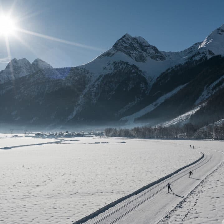 Drei Personen fahren auf einer schneebedeckten Piste in einem weiten Tal, mit hohen, sonnenbeschienenen Bergen im Hintergrund unter einem klaren blauen Himmel.