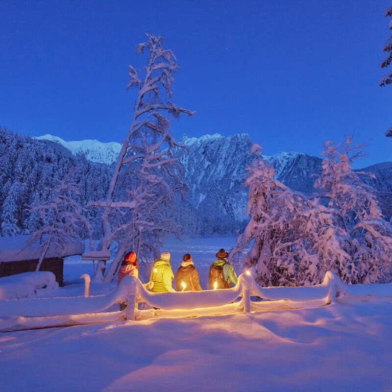 Vier Personen in Winterkleidung stehen in der Abenddämmerung an einem verschneiten Zaun, umgeben von schneebedeckten Bäumen und Bergen, die von warmen Lichtern beleuchtet werden.