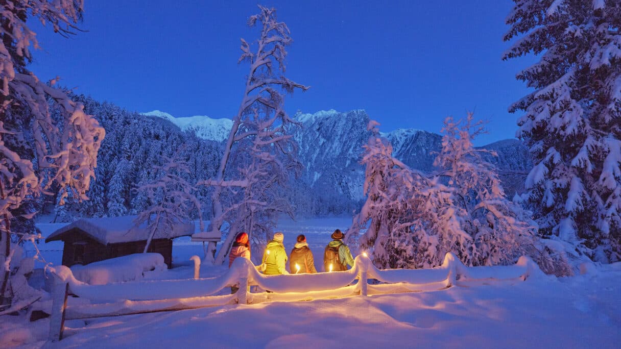 Drei Personen in Winterkleidung stehen in der Abenddämmerung an einem verschneiten Zaun und halten Laternen, umgeben von schneebedeckten Bäumen und Bergen.