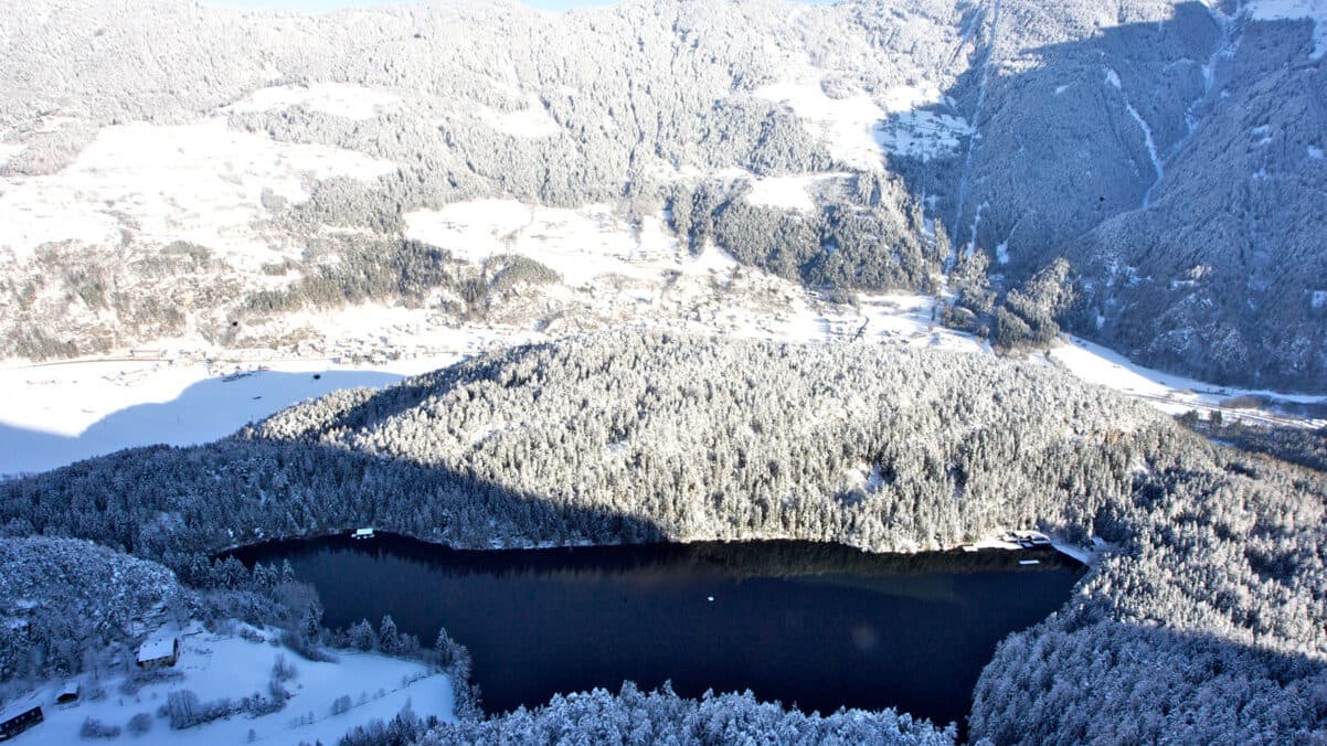 Luftaufnahme einer schneebedeckten Berglandschaft mit einem dunklen, nicht zugefrorenen See, umgeben von Bäumen und verstreuten Gebäuden.