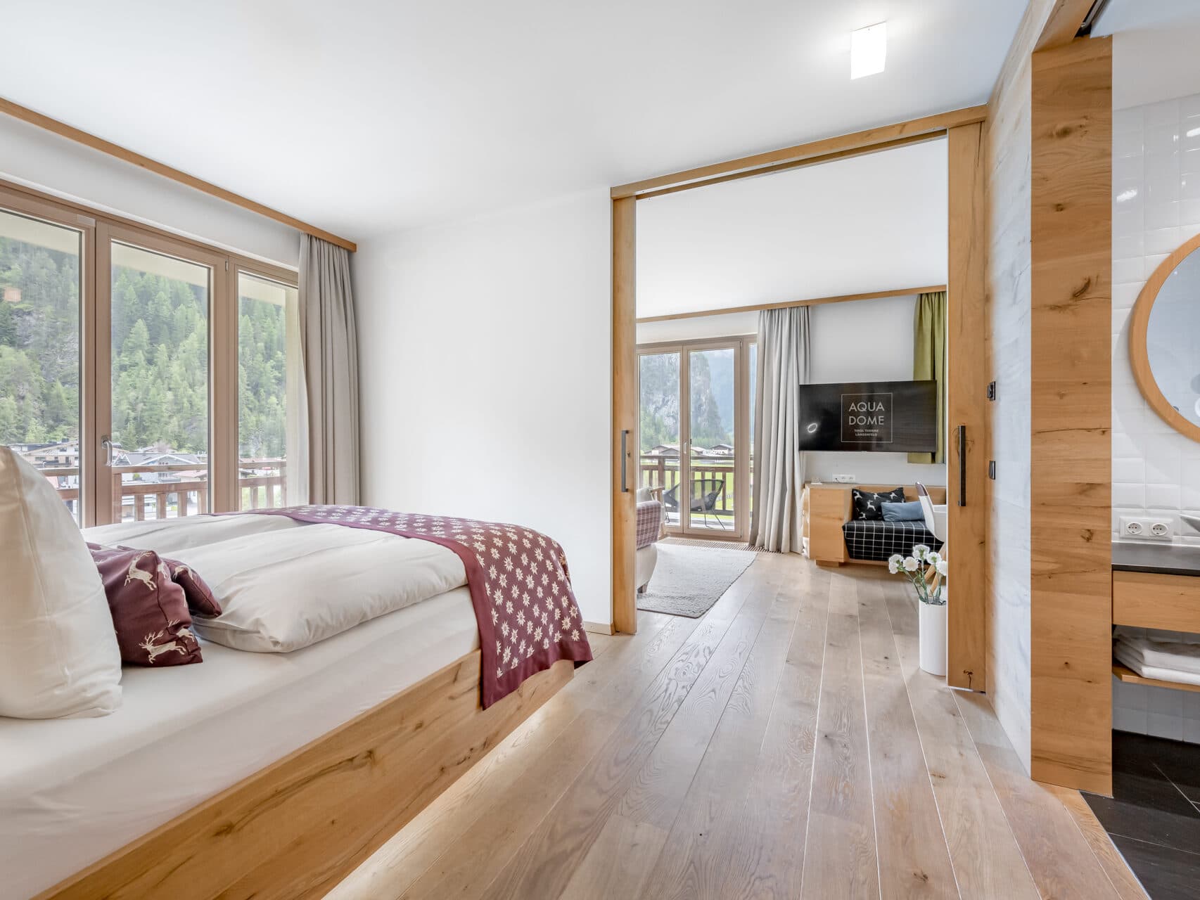 Moderne Hotelsuite mit Doppelbett, Holzboden, Schiebetüren, Balkon, Sitzecke mit TV, großen Fenstern und eigenem Bad.