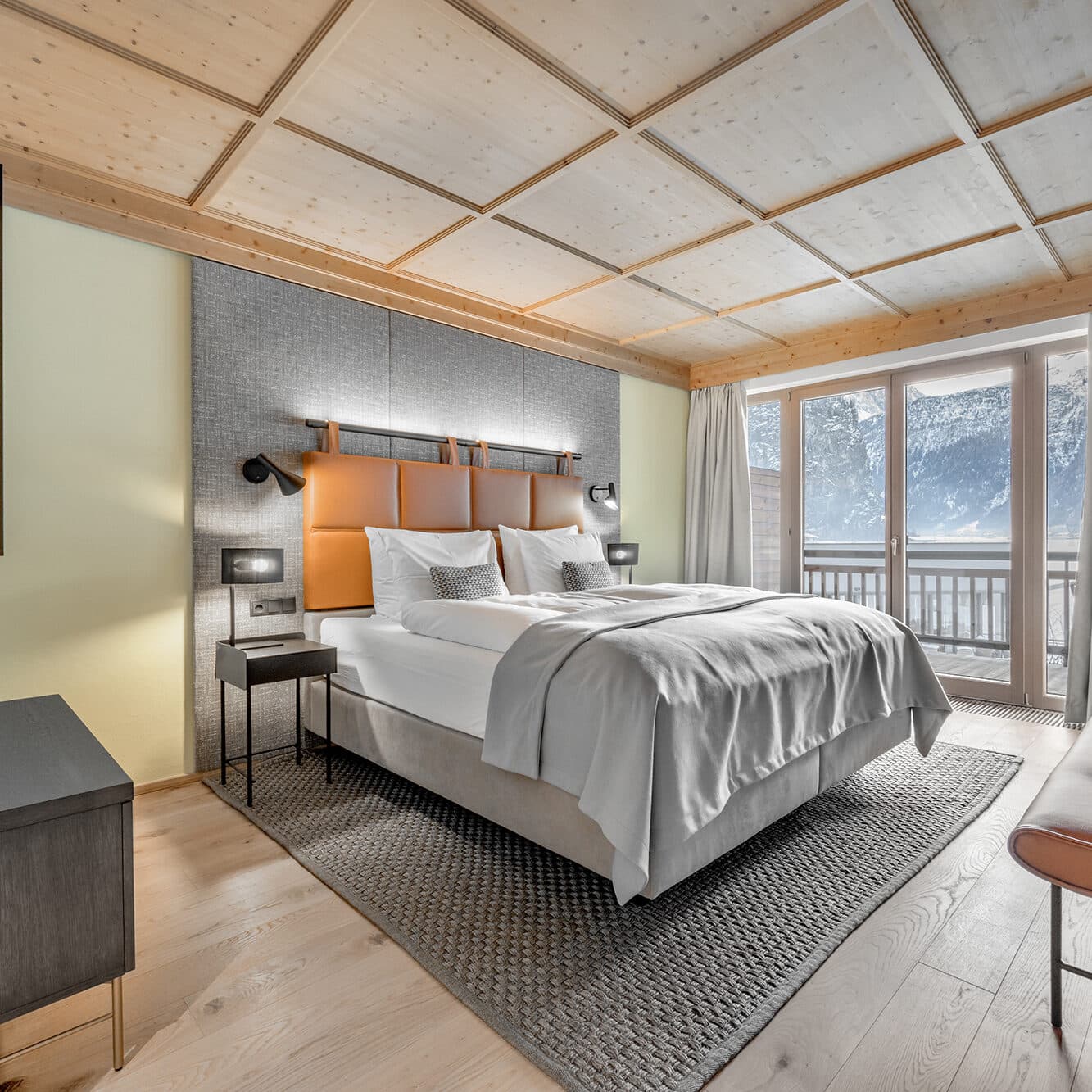 Modernes Hotelzimmer mit großem Bett, Nachttischlampen, Holzdecke und -boden, Teppich und Fenster mit Blick auf die Berge.