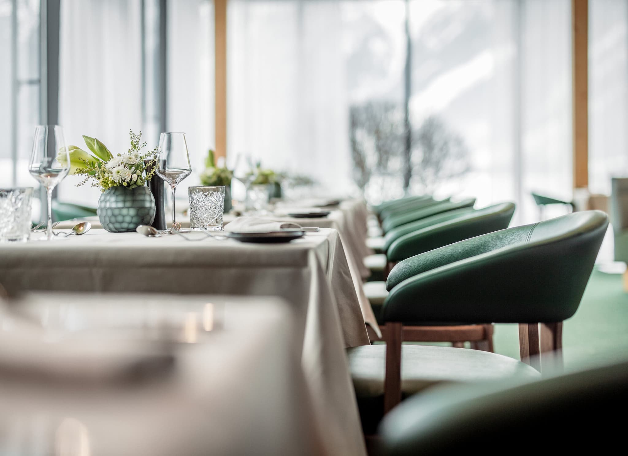 Ein modernes Restaurant mit ordentlich gedeckten Tischen, grünen Stühlen, Glaswaren und Blumenschmuck, mit großen Fenstern im Hintergrund.