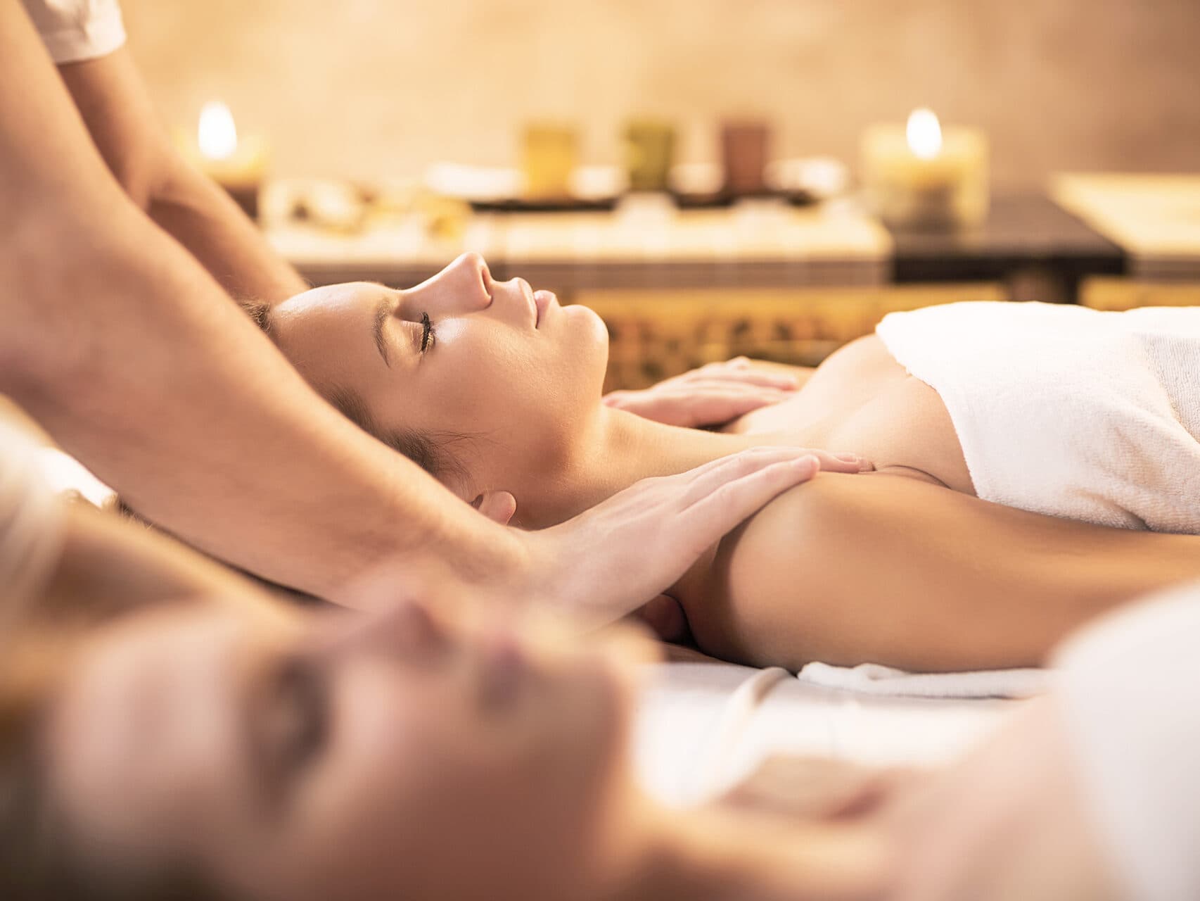 Zwei liegende Personen erhalten eine Nacken- und Schultermassage in einer Spa-Umgebung mit Kerzen und sanfter Beleuchtung im Hintergrund.