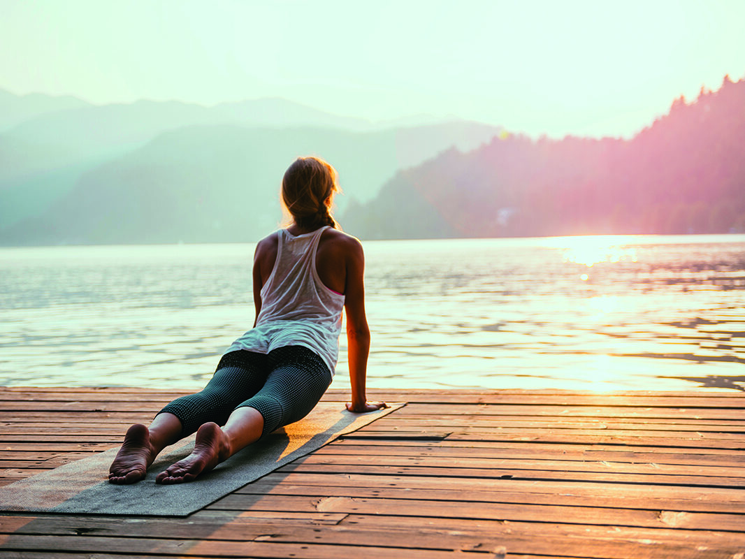 Eine Person, die auf einer Matte auf einem hölzernen Steg an einem See bei Sonnenaufgang oder Sonnenuntergang Yoga in einer nach oben gerichteten Hundestellung praktiziert.