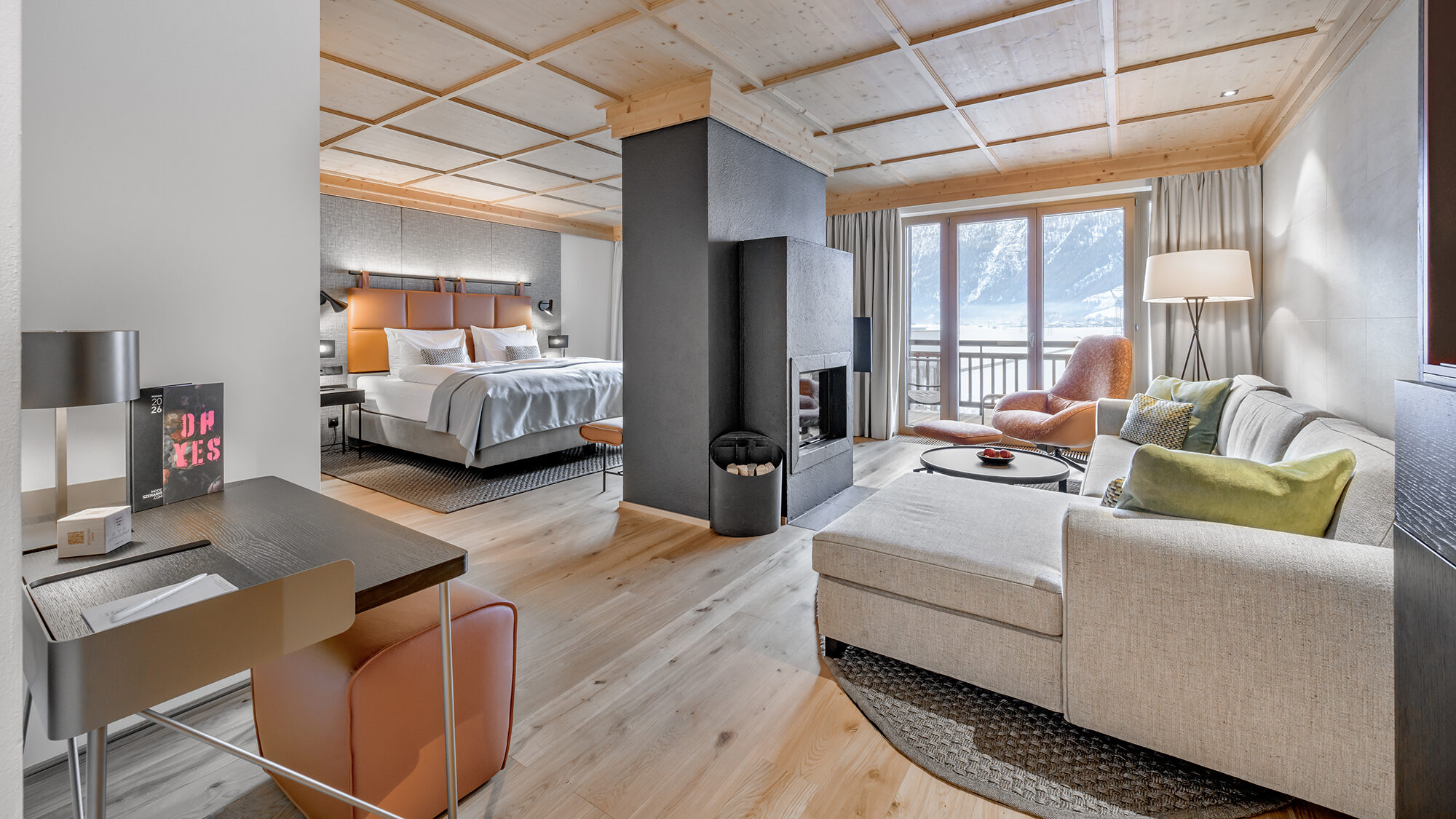 Modern hotel suite with a king bed, desk, fireplace, seating area, and large window offering a mountain view. Light wood floors and neutral-toned furnishings create a cozy atmosphere.