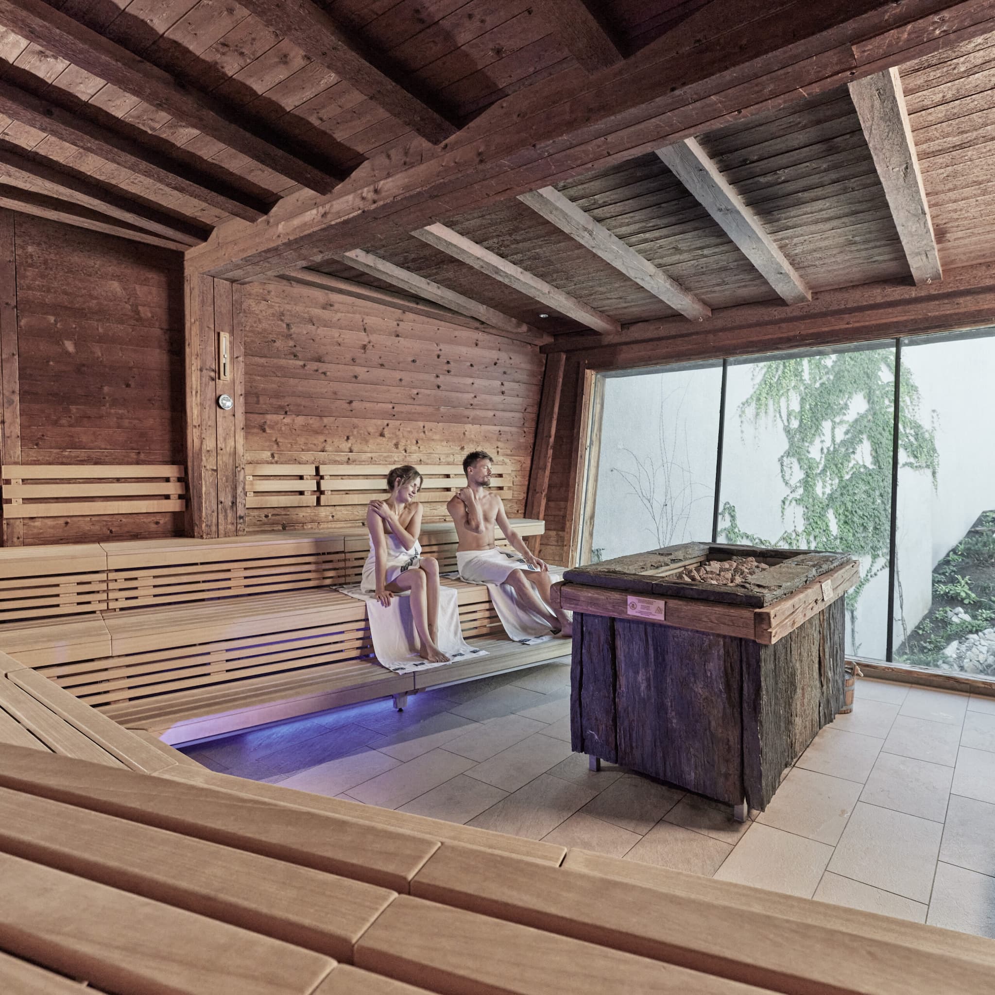 Two people sit on wooden benches in a modern sauna room, wrapped in towels, with a large window showing plants outside.