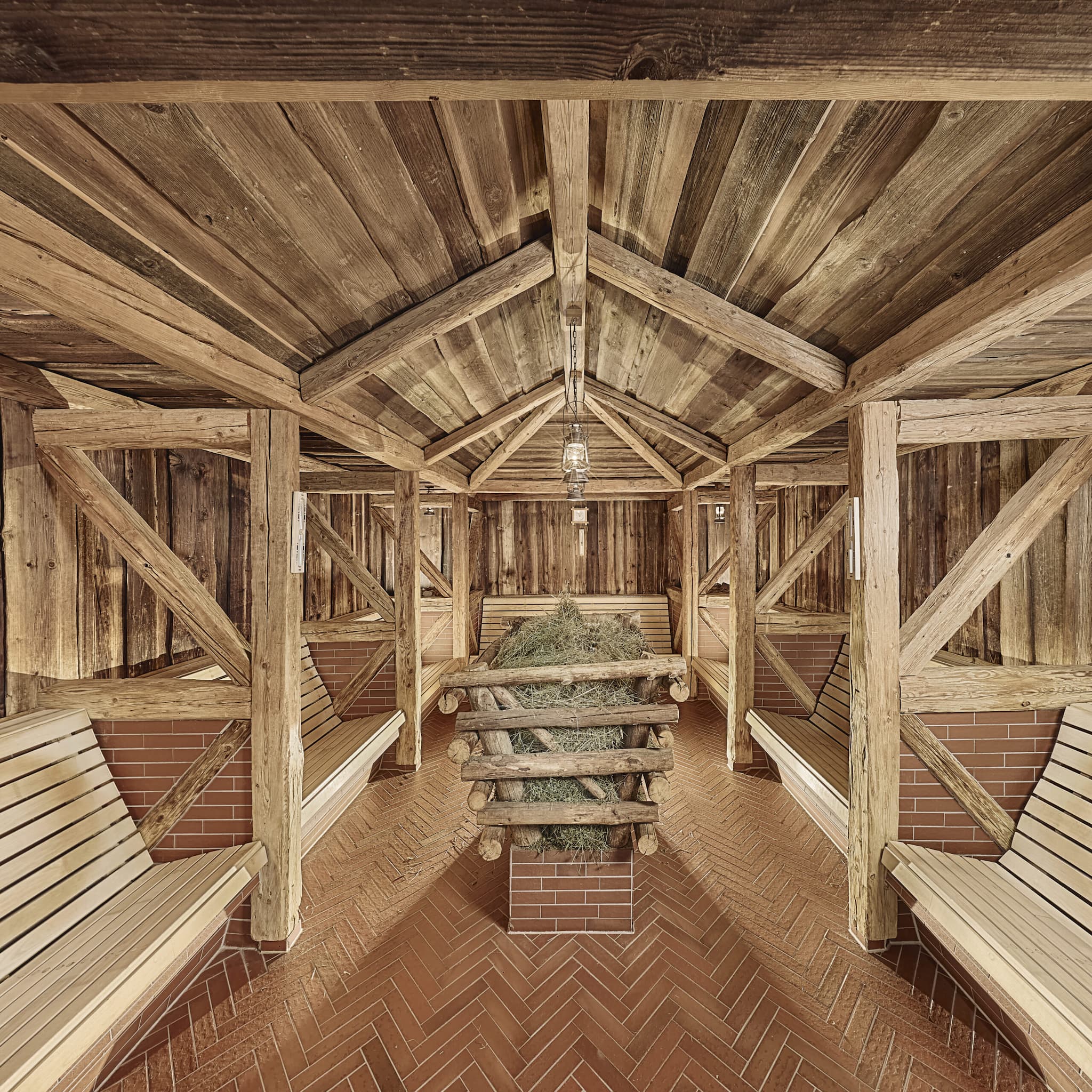 Wooden sauna room with angled benches, exposed beams, brick floor, and a central structure filled with hay under a hanging light.